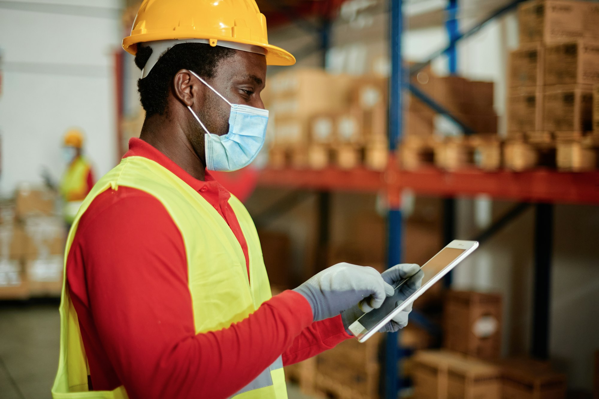 Caucasian adult warehouse worker checks delivery order on a tablet while wearing a safety mask